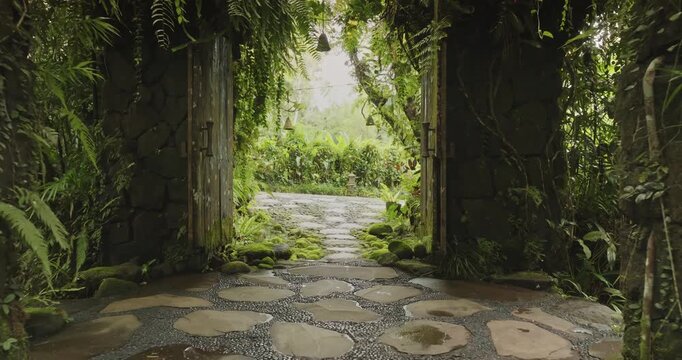 Travel to Bali. Ancient stone doorway providing a tranquil entrance to a lush tropical garden featuring a moss-covered path, abundant green foliage, ferns, and hanging bells for a serene experience