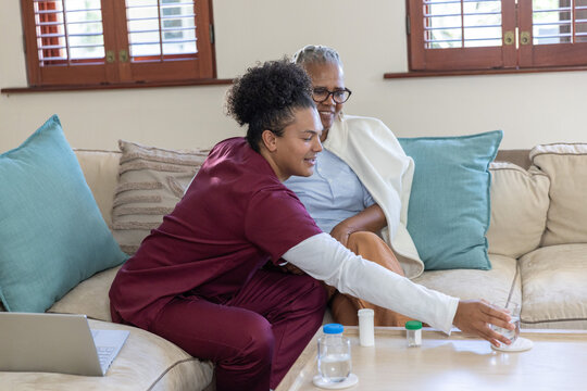 Senior African American woman and aide sitting on sofa placing water near meds in burgundy scrubs