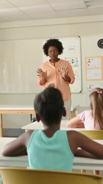 Gesturing teacher wearing peach blouse speaking to students at school, with board, posters, clock