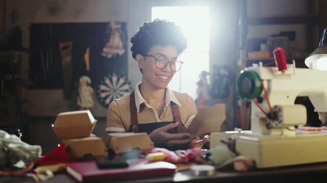 A person makes dolls from fabric pieces in a well-lit workshop. The workspace is filled with materials and tools. The person smiles while working on the craft.