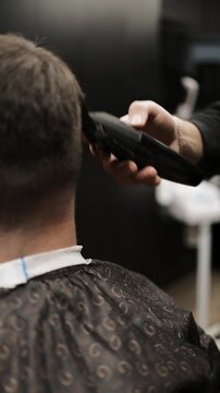Close-up of professional barber trimming a client's hair with electric clipper. Back view of male grooming process with stylish patterned cape and neck strip in barbershop.