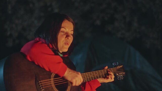 Intimate singer with campfire melody. Serene scene of musician playing guitar by flickering campfire. Calm evening atmosphere with musician strumming guitar near tent under warm firelight