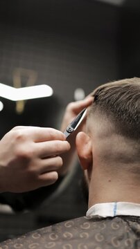 Close-up of professional barber trimming a client's hair with electric clipper. Back view of male grooming process with stylish patterned cape and neck strip in barbershop.