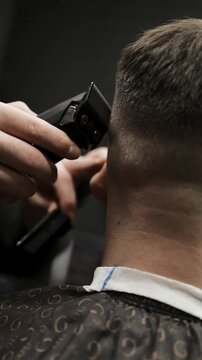 Close-up of professional barber trimming a client's hair with electric clipper. Back view of male grooming process with stylish patterned cape and neck strip in barbershop.