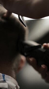 Close-up of professional barber trimming a client's hair with electric clipper. Back view of male grooming process with stylish patterned cape and neck strip in barbershop.