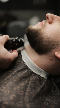 Close-up of professional barber trimming a client's beard with electric clipper and comb. Side view of male grooming process with patterned cape and neck strip in barbershop.