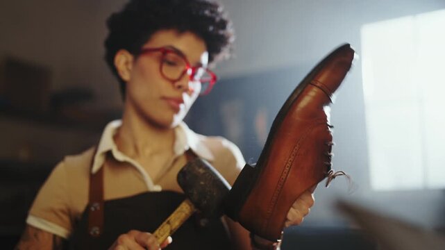 A woman with curly hair and glasses is focused on crafting a leather item in a workshop. Tools and materials are around her as she pays attention to her work.