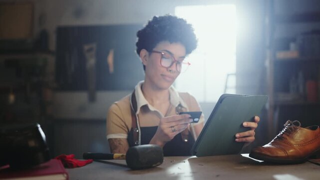A person sits at a table in a workshop. They hold a tablet and a card while looking focused. Tools and a shoe are on the table. Sunlight comes through a window.