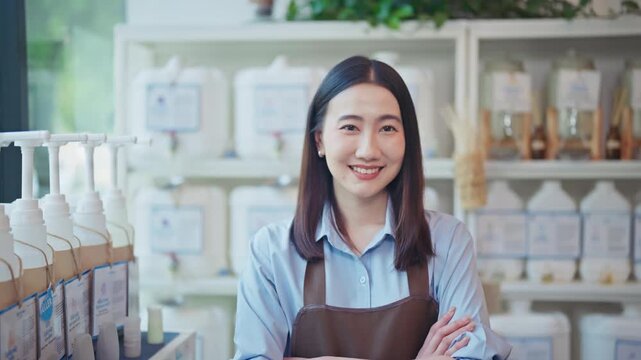 Proud young Asian woman in an apron stands confidently with arms crossed and a bright smile inside her clean zero waste organic refill shop filled with natural products.