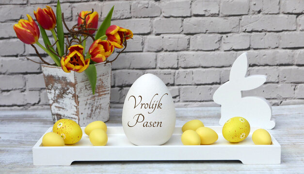 Dutch Easter greeting: 'Vrolijk Pasen' on a white egg with yellow eggs, tulips and wooden rabbits in front of a brick wall.