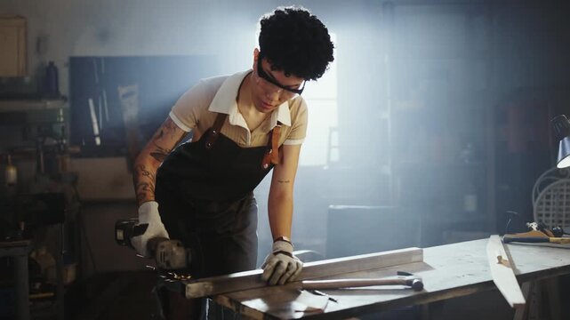 An urban woman uses a grinder tool to work on metal pieces in a workshop. The light from a window creates a bright effect. Tools and materials are spread across the table.