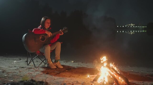 Lakeside acoustic guitarist playing by campfire, soft firelight and gentle strums, smoke drifting over water, distant lights on opposite shore, folding chair and hoodie, cozy boots on sand, intimate