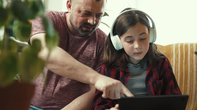 Preteen girl in headphones showing interesting content on digital tablet to her father at home-4k,10 bit footage.