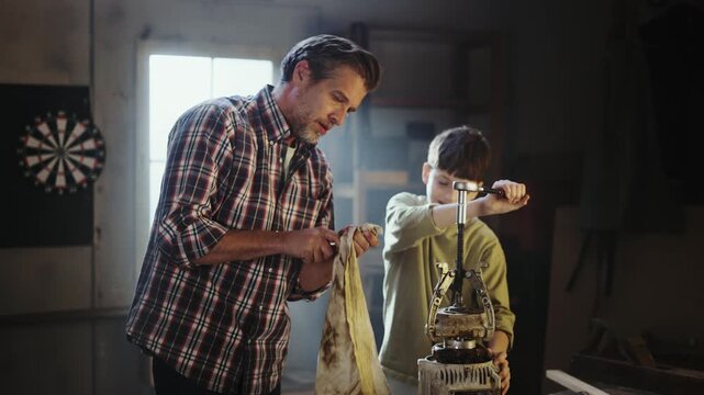 A father helps his son as they work on a project in a workshop. They focus on a kettle while using tools. Light shines through the windows creating a bright space.