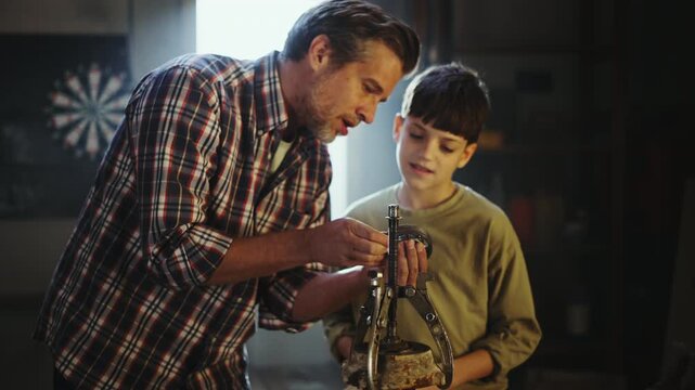 A father helps his son build something new in their workshop. They share ideas as they focus on the task. The space is bright with natural light coming through a window.