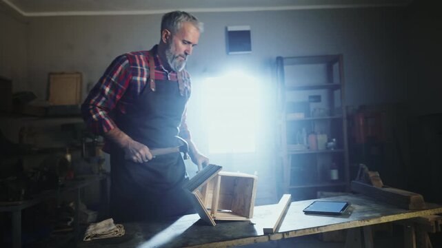 A man works in a dim workshop. He uses a hammer to build a wooden box. Tools and wood are around him. Light shines down, showing his focus on the task.