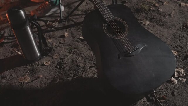 Black guitar on sandy ground, camping still life with thermos, dim ember glow, closeup of strings and frets, soft shadows, moody nighttime atmosphere, empty campsite vibe, rustic texture and warmth