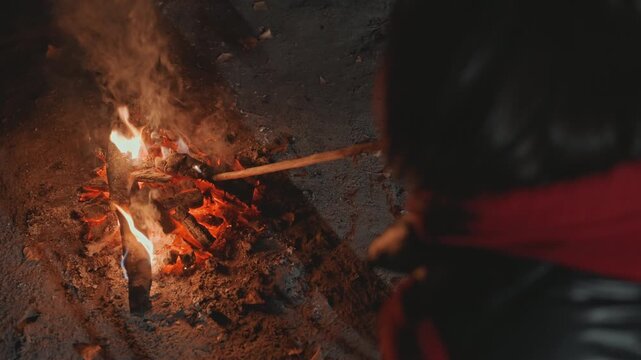 Night camping scene with campfire, camper in red scarf stokes glowing embers with stick while roasting marshmallow, leather jacket and hands visible, ash and sparks rising into smoky air, intimate
