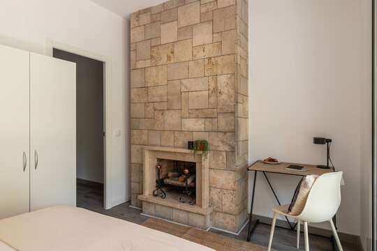 A modern bedroom featuring a stone fireplace, white wardrobe, and a desk with a chair. The room has neutral colors and a minimalist design.