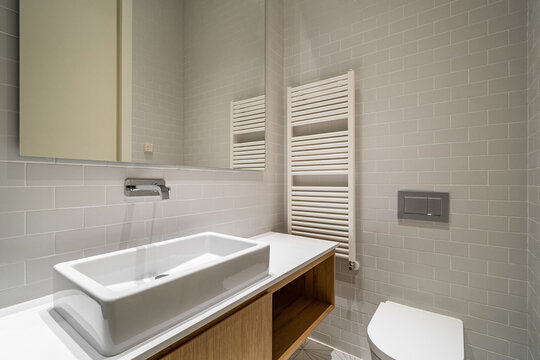 Modern bathroom with a sleek design. Features a white sink, wooden cabinet, and a towel radiator. Neutral color palette with gray tiles and a large mirror.