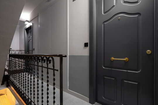 A modern hallway with gray walls and a black railing. A closed door with a brass handle is visible. The floor is tiled and well-lit.