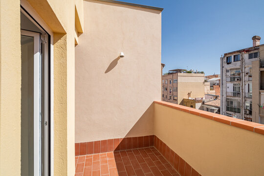 A sunny balcony with a terracotta floor and beige walls. The view includes nearby buildings under a clear blue sky.