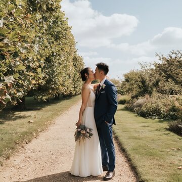 Bride female asian young and groom male caucasian adult kissing on sunlit garden path wedding dress bouquet trees greenery romantic moment
