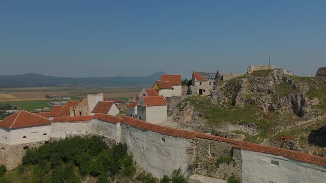 Extensive aerial pull-out over the historic Rasnov Citadel revealing newly reconstructed medieval houses alongside ancient ruins under a clear blue sky, graded