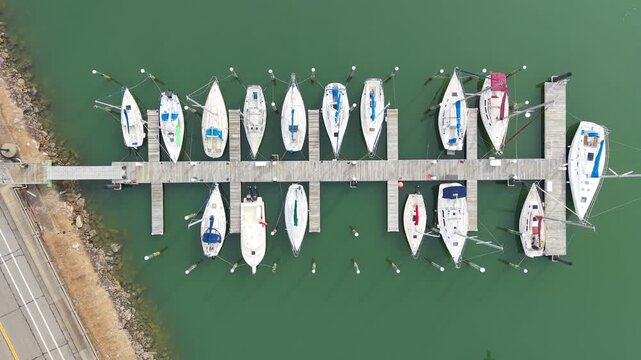 Aerial top-down view of various sailboats and motorboats docked in wooden slips at a calm coastal marina during the day.