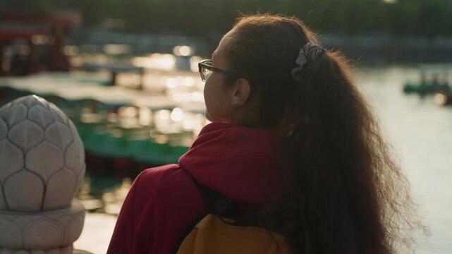 Young woman gazing at golden water, soft evening light highlights ponytail and glasses, backpack on shoulders, boats and marina shimmer, urban skyline blurred, contemplative mood, cinematic side