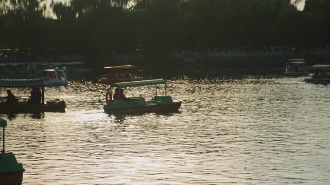 Sunlit lake with paddle boats gliding golden evening light reflects off water as families and couples enjoy leisurely pedal boating near treelined shore and busy marina, creating peaceful urban