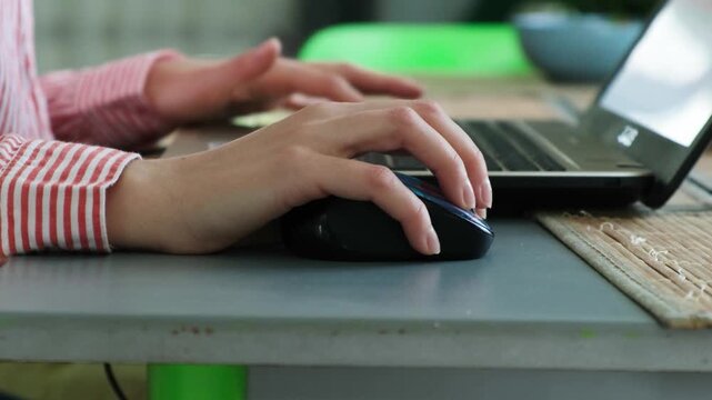 Close-up view of a female freelancer's hands using a wireless computer mouse and typing on a laptop keyboard, engaged in remote work or online browsing from a home office environment