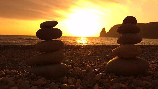 Zen sunset beach, two balanced stone stacks on a pebble beach at a serene sunset with ocean and distant hills.