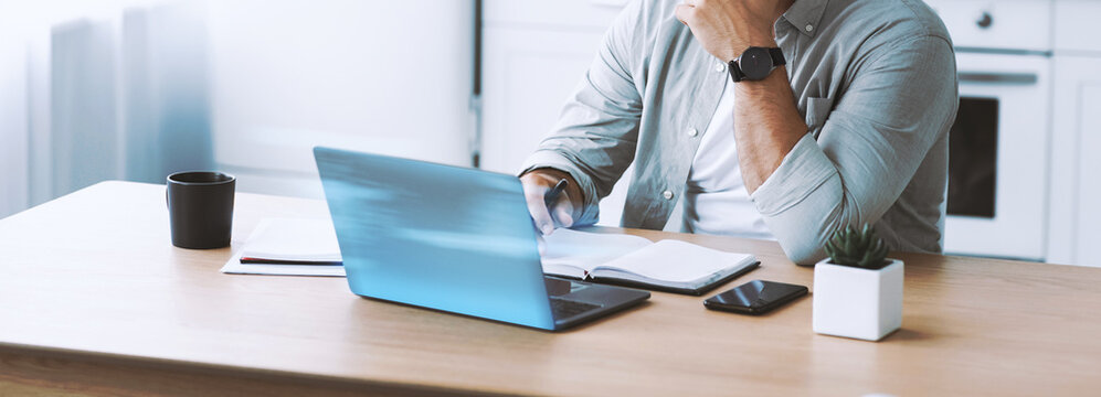 Middle aged european businessman in glasses works on laptop at kitchen table. He is focused on business tasks, managing bills and continuing education during covid-19.