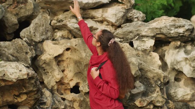Caucasian woman in red jacket examines textured stone up close, delicate hand tracing crevices, naturalist and geologist persona, contemplative focus, soft daylight, green park backdrop.