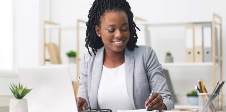 Black Millennial Businesswoman Taking Notes And Using Laptop While Working In Modern Office. Horizontal Banner, Panorama With Free Space