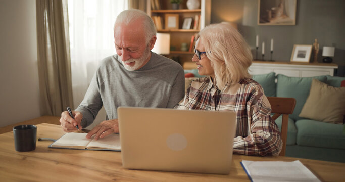 Elderly couple sits at a table with a laptop open, discussing life insurance. One person takes notes while the other looks at documents. They appear engaged in financial planning.