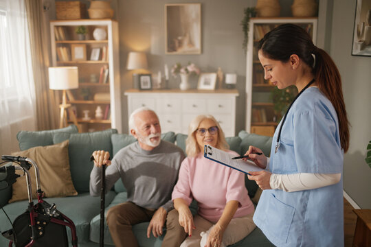 Home health nurse stands with clipboard in hand as she talks to an elderly couple sitting on the couch in their living room during a care visit