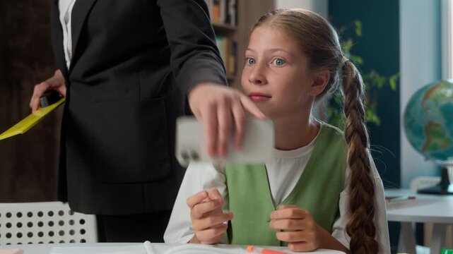 Caucasian little girl child kid pupil playing video game with mobile phone at class lesson in school unrecognizable woman teacher female taking away smartphone control education discipline behavior