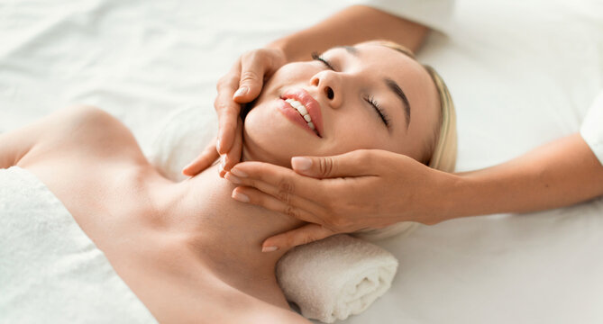 Young Blonde Woman Enjoying Relaxing Massage Lying On Bed, Masseur Massaging Her Neck And Face At Spa Indoors, Closeup. Relaxation, Beauty And Wellness Concept. Panorama