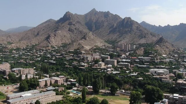 Aerial drone view of a city located in a deep valley surrounded by tall mountains. A river runs through the town center on a cloudy day in summer.