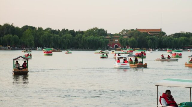 Sunset lake crowded with pedal boats, families and friends aboard colorful rental vessels, gentle ripples across calm water, green treeline and distant pavilion, warm pastel sky creating relaxed urban