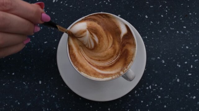 Top view of hand with pink nails stirring frothy cappuccino coffee