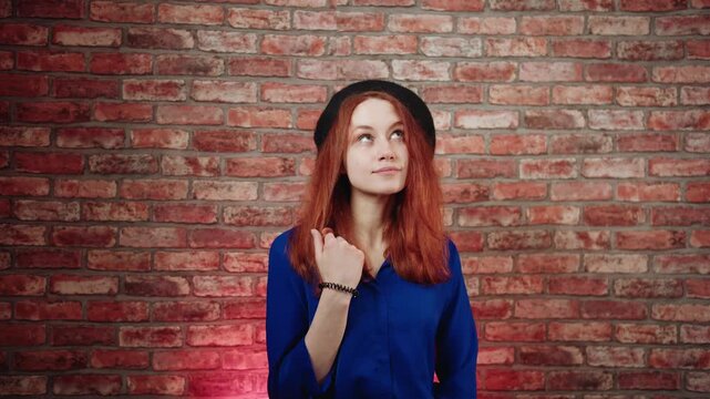 A portrait of a dreaming joyful young caucasian redhead woman in a blue blouse and black beret, fantasizes and thinks about pleasant good things against a brick wall. She twirls a hair and looks up