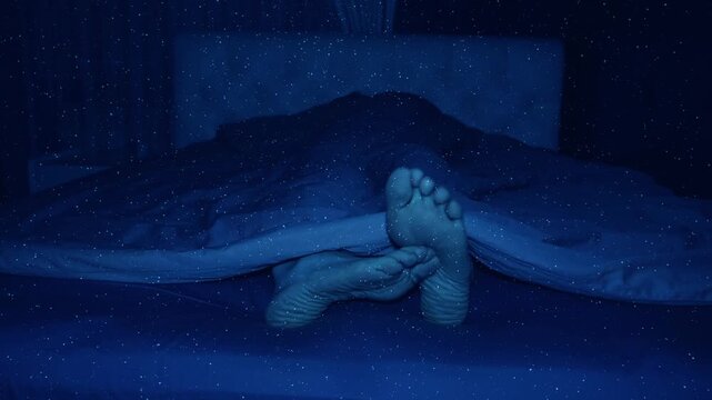 In the darkness of the bedroom bed, a man sleeps soundly, only occasionally one foot lazily scratches the other foot. View through sparkling particles. Shot in motion