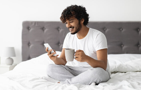 Cheerful handsome dark-skinned man in pajamas sitting on bed at home, drinking coffee and using smartphone, checking social media while chilling alone at weekend, copy space. Leisure and gadgets