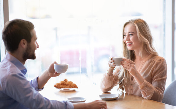 Portrait of cute affectionate couple sitting at table in city cafe, drinking aromatic coffee, enjoying each other's company. Cheerful man with his girlfriend having friendly chat on date at restaurant