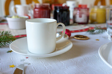 Fototapeta premium white ceramic mug on saucer with Christmas decorations and jam jars in background, festive holiday setting