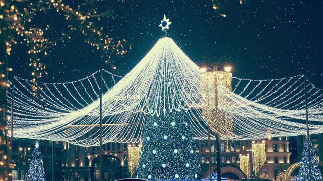 Beautiful, like in a fairy tale, richly decorated Christmas tree with luminous garlands and balls in the city square creates a festive, New Year's mood. View through sparkling particles. Shot in motio