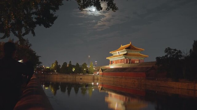 Moonlit pavilion by moat silhouetted photographer capturing ornate tower reflection under clouded sky, warm lantern glow, trees framing walkway, calm water mirror, contemplative nocturnal atmosphere,
